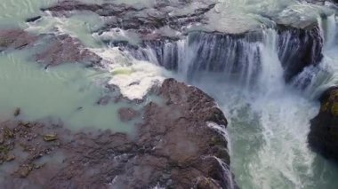 Volkanik arazi, yosunlu yeşillik ve seyrek bitki örtüsüyle çevrili turkuaz bir nehre akan çok katmanlı bir şelalenin havadan görünüşü..