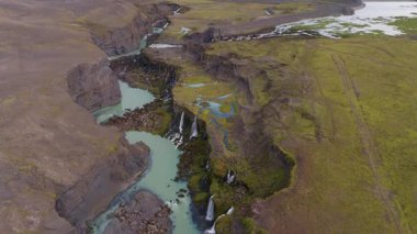 Drone, İzlanda 'da küçük şelaleleri, volkanik oluşumları ve canlı yeşil yosunları olan kayalık bir kanyonda süzülen bir turkuaz nehri yakalar..