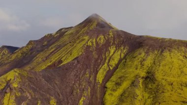 İnsansız hava aracı İzlanda 'da volkanik bir dağın etrafında yükselir, koyu kayalar, yeşil yosunlar, sis ve çevresindeki ıssız bir araziyi gösterir..