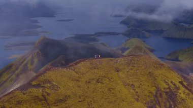 İzlanda 'da dar bir tepe boyunca yürüyen, yosun kaplı arazi ve karanlık volkanik toprak ile çevrili iki yürüyüşçünün hava görüntüsü..