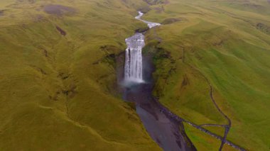 İzlanda 'daki Skogafoss şelalesinin hava görüntüleri çağlayanı, sisi, yeşil tepeleri, dolambaçlı bir nehri, park alanını ve yürüyüş yollarını gösteriyor..