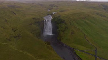 İzlanda 'daki Skogafoss şelalesinin hava manzarası, çağlayan su, sisli havuz, yemyeşil tepeler, görünür yollar ve küçük ziyaretçi figürleri..