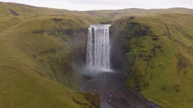 İzlanda 'daki Skogafoss şelalesinin hava manzarası, güçlü çağlayanı, sisli havuzu ve bulutlu gökyüzünün altındaki yemyeşil tepeleri gösteriyor..