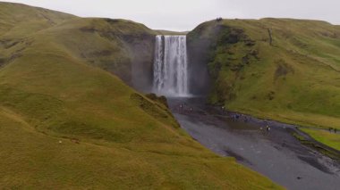 İzlanda 'daki Skogafoss şelalesinin hava görüntüsü, çağlayan suyu, sisi, yeşil tepeleri, volkanik araziyi ve ziyaretçi aktivitesi olan patikaları gösteriyor..