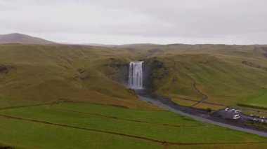 İzlanda 'daki Skogafoss şelalesinin hava manzarası sisli, yemyeşil tepeler, akan bir nehir, park edilmiş arabalar ve kırmızı çatılı bir bina..