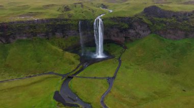 İzlanda 'daki Seljalandsfoss şelalesinin hava manzarası, bir havuza dökülen su, yemyeşil alanlar, yürüyüş yolları ve nazik insan aktiviteleri..