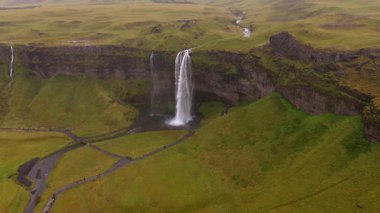 Seljalandsfoss şelalesinin yeşil tarlalar, yürüyüş yolları ve İzlanda 'daki küçük insan gruplarıyla çevrili bir havuza düşüşünün havadan görüntüsü..