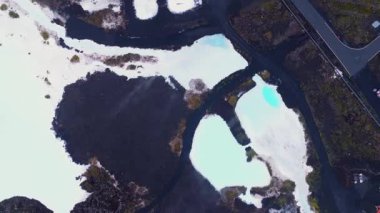 İzlanda 'nın Mavi Göl' ünde pürüzsüz bir insansız hava aracı uçuşu. Süt mavisi jeotermal havuzlar, karanlık volkanik kayalar ve sağ tarafta bir yol sergiliyor..