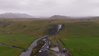 İzlanda 'daki Skogafoss şelalesinin havadan görünüşü, bir nehre dökülen su, yemyeşil tepeler, tarım arazileri ve bulutlu bir gökyüzünün altındaki park alanı..