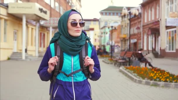 Uma turista menina em um lenço de cabeça e óculos de sol em uma mochila andando pela rua — Vídeo de Stock