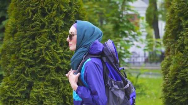 Seorang gadis turis dengan kerudung dan kacamata hitam dengan ransel berjalan di taman — Stok Video