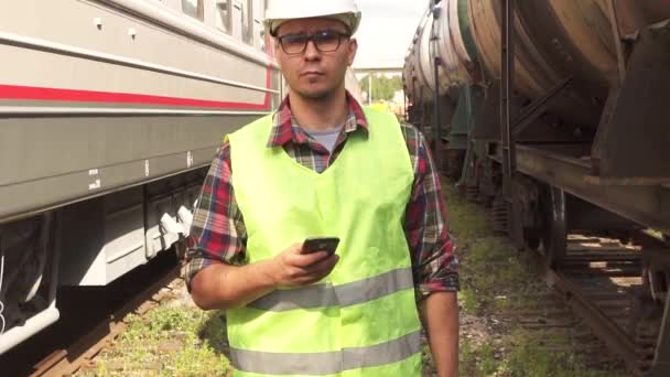 Retrato de un hombre en gafas trabajador en un casco y uniforme, caminando por la estación de tren con el teléfono — Vídeos de Stock