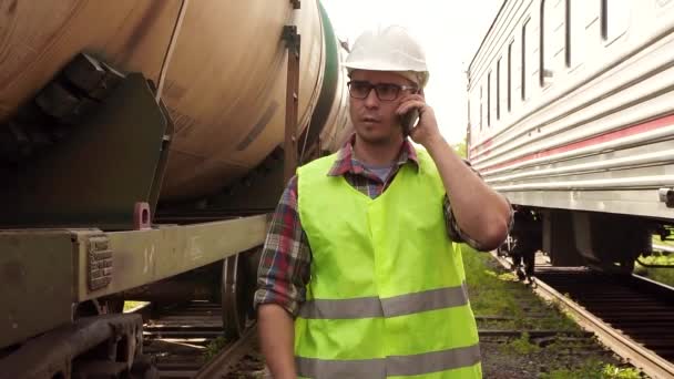 Портрет человека в очках рабочего в шлеме и униформе, идущего по вокзалу, разговаривающего по телефону — стоковое видео