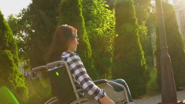 Девушка-инвалид в футболке в инвалидной коляске на закате проезжает по парку. — стоковое видео