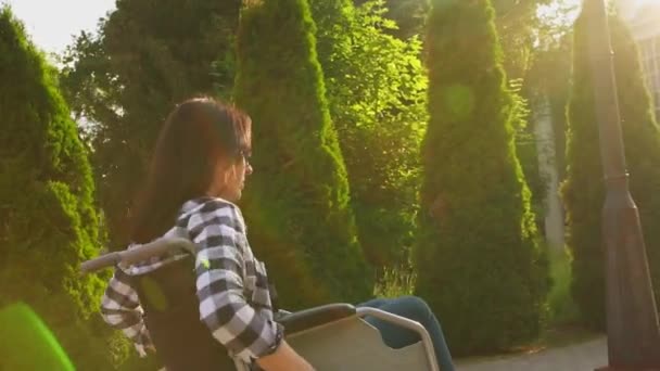 Funktionshindrade flicka i en skjorta i rullstol rider genom parken vid solnedgången — Stockvideo