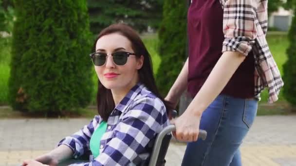 Девушка в рубашке закатывает девочку-инвалида в очках и рубашку в инвалидном кресле. И смотрит в камеру — стоковое видео