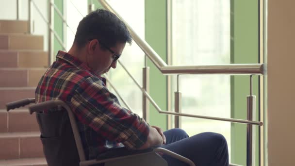 Одинокий инвалид в очках и рубашке в инвалидном кресле смотрит в окно и грустно — стоковое видео