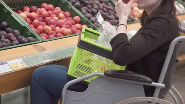 Una mujer con una discapacidad en una silla de ruedas de compras en el supermercado elige frutas y las pone en un paquete. — Vídeo de stock