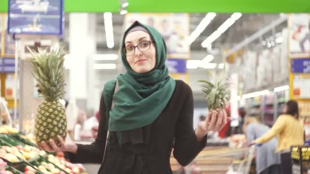 Portret van een moslim meisje in hijab in een supermarkt die in het bezit van de ananas. Weet niet welke te kiezen — Stockvideo