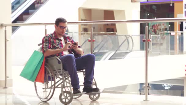 Un uomo disabile su una sedia a rotelle che usa il telefono al centro commerciale dopo aver fatto shopping — Video Stock