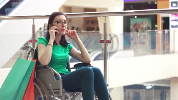 Una mujer con una discapacidad en una silla de ruedas se divierte hablando por teléfono en el centro comercial después de comprar. — Vídeo de stock