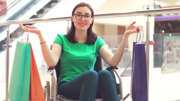 Una donna disabile su una sedia a rotelle mostra pacchetti per lo shopping e sorride alla macchina fotografica. — Video Stock