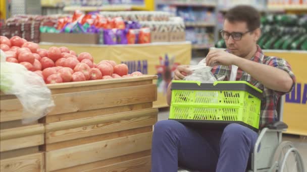 Человек с инвалидностью в инвалидной коляске, покупающий в супермаркете, выбирает помидоры и кладет их в упаковку. — стоковое видео