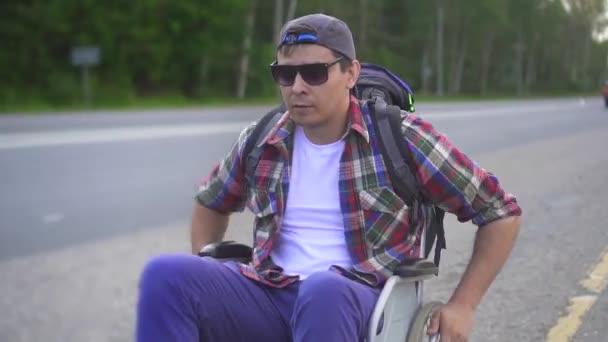 Человек с инвалидностью в инвалидном кресле с рюкзаком, путешествующим по шоссе. Закрыть — стоковое видео
