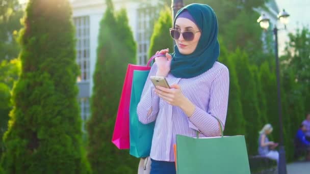 Jeune femme musulmane dans des sacs de hijab dans ses mains après avoir fait des achats en utilisant le téléphone sunflare.Slow mo — Video