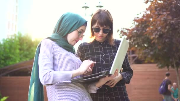 Studentessa musulmana in hijab e la sua amica con computer portatile in chat nel parco, sunflare . — Video Stock