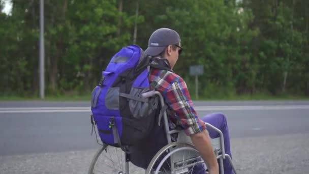 Człowiek z osobą niepełnosprawną na wózku inwalidzkim z plecakiem podróży autostradą. Widok z tyłu — Wideo stockowe