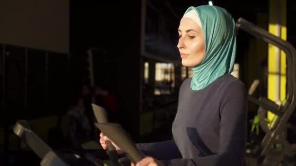 Jovem mulher muçulmana esportiva no hijab fazendo esportes no simulador na frente da janela.Fechar — Vídeo de Stock