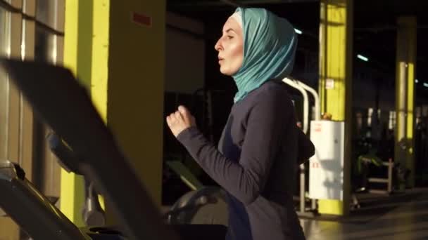 Портрет спортивной мусульманки в шарфе на тренажере в тренажерном зале — стоковое видео