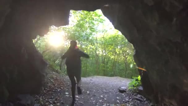 Una turista con una mochila y una linterna en la frente sale corriendo de la cueva oscura y levanta las manos, rayo de sol — Vídeo de stock