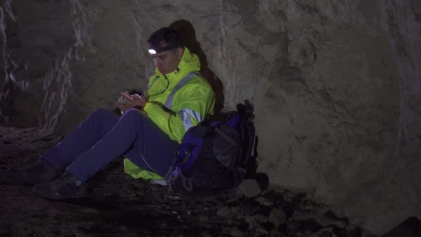 Turista hombre con una linterna en la frente y un teléfono en la mano perdido en la cueva, sentado y tratando de llamar a los rescatistas — Vídeos de Stock