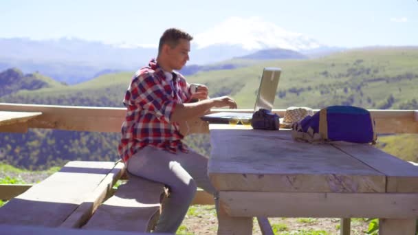 Людина є позаштатним працюють на ноутбук і пити з чашки на тлі гір — стокове відео