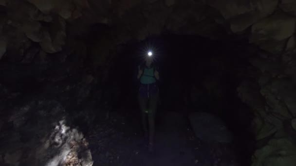 Flicka turist med en ryggsäck och en ficklampa i pannan går ut ur den mörka grottan — Stockvideo