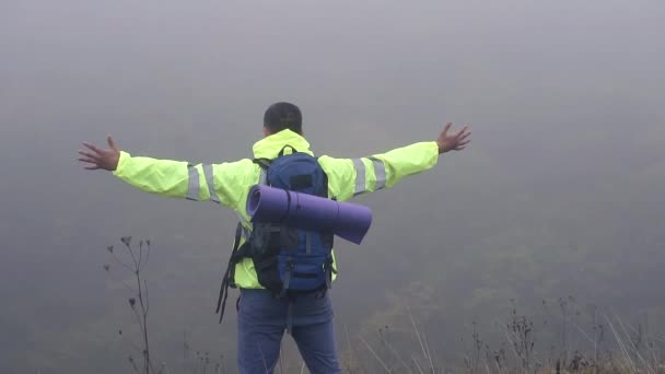 Человек турист с рюкзаком стоит на краю горы в тумане размахивая руками, медленно мо — стоковое видео