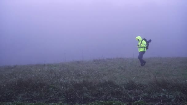 Турист в сигнальном жилете в капюшоне с рюкзаком, ходящим в тумане. — стоковое видео