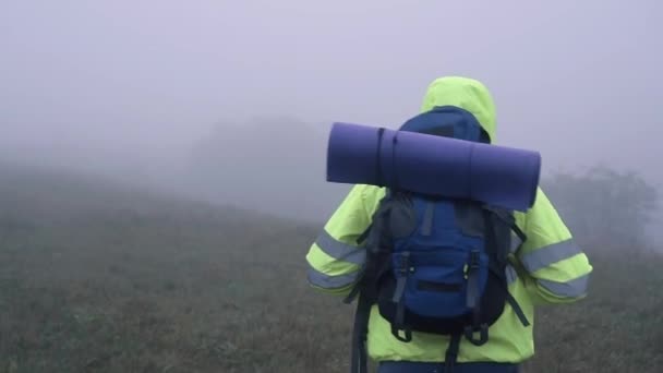 Портрет туриста-чоловіка в сигнальному жилеті в капюшоні з рюкзаком, що йде туманним видом ззаду — стокове відео