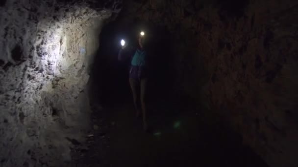 Ragazza turista con uno zaino con una torcia elettrica sulla fronte e nelle mani è su una caverna scura ed esplorarlo — Video Stock