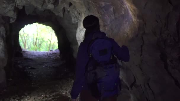 Ragazza turista con uno zaino va alla grotta con una torcia sulla fronte ed esamina le pietre, vista posteriore — Video Stock