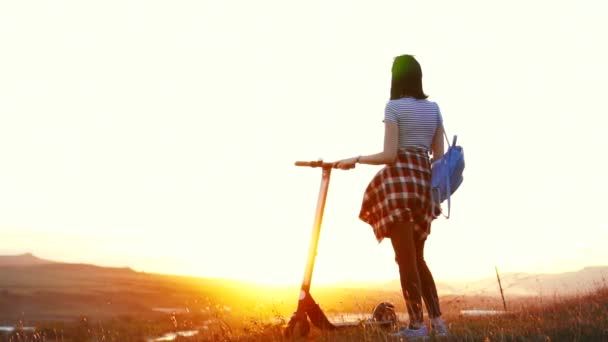 Chica con un scooter eléctrico se para con su espalda y mira a la puesta de sol, lento mo — Vídeos de Stock