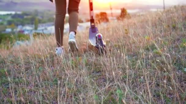 Mädchen Beine geht und rollt den Elektroroller neben der Sonnenuntergangssonne, langsam mo Nahaufnahme — Stockvideo