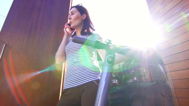 Ung flicka talar på en smartphone och håller en hand på elektrisk skoter, solen sken — Stockvideo