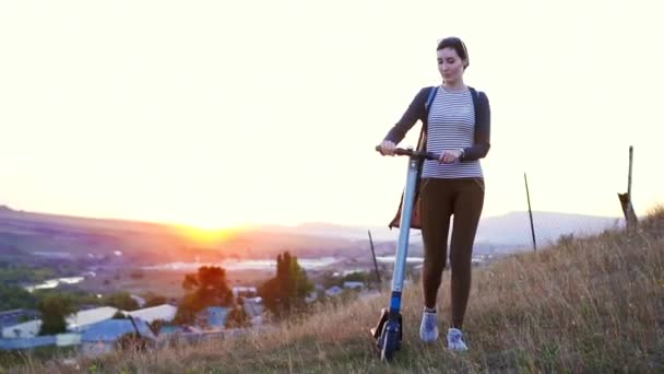 Chica joven camina por la colina con un scooter eléctrico y observa la puesta de sol . — Vídeos de Stock