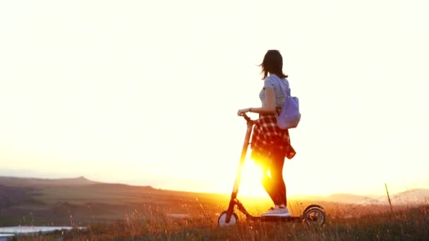 Chica con un scooter eléctrico de pie con la espalda mirando a la puesta de sol y levanta la mano con alegría, lento mo — Vídeo de stock