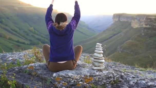 Mädchen sitzt in Lotusposition auf einem Felsen am Berg bei Sonnenuntergang, Rückansicht. das Konzept der Meditation und der Natur — Stockvideo