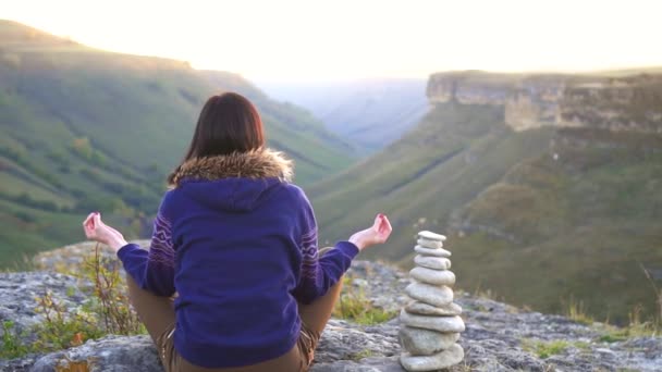 Fille assise en position lotus au sommet d'une montagne au coucher du soleil, vue arrière — Video