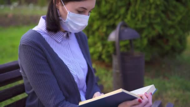 Retrato de una niña con una máscara médica protectora en la cara y con un libro en un banco al atardecer — Vídeo de stock